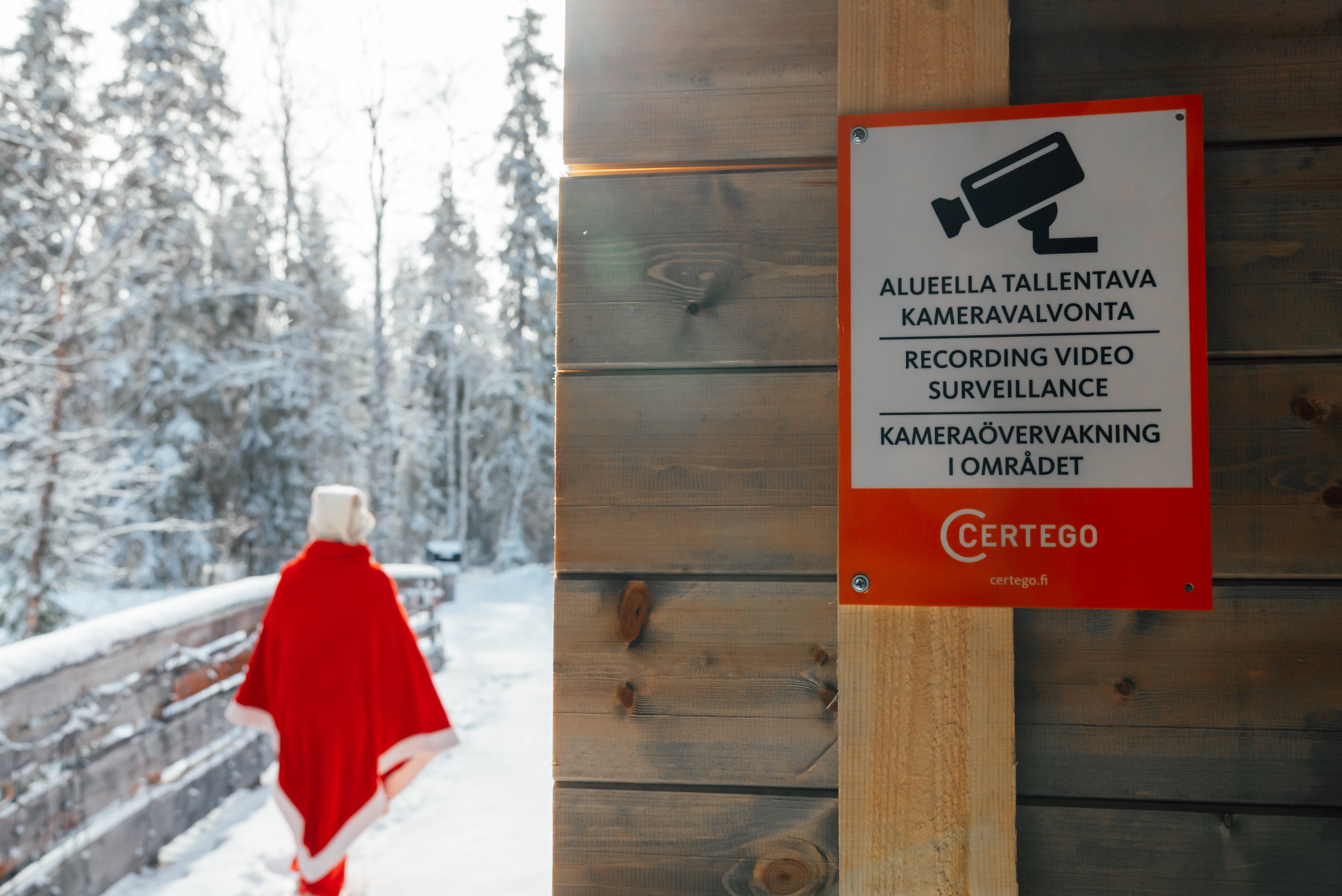 Tonttu kävelee elämyskylän portista läpi, taustalla CERTEGOn kameravalvontakyltti