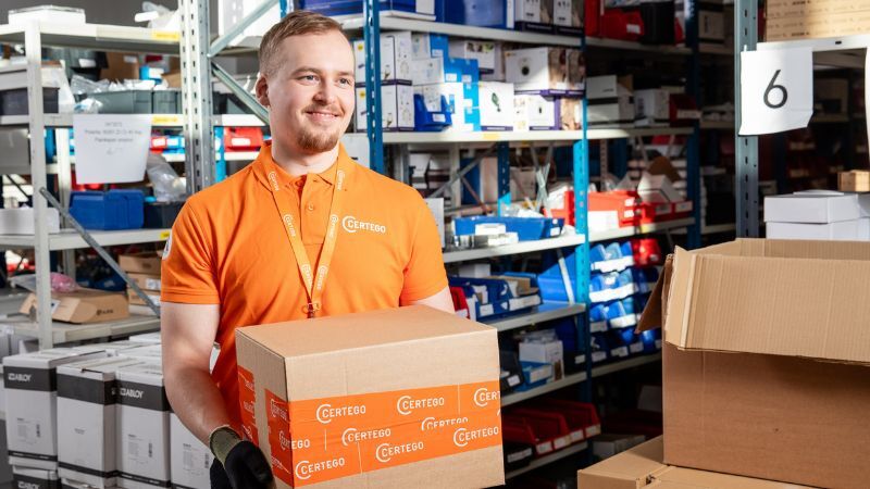 Varastopäällikkö pitää certego teipattua pahvilaatikkoa kädessään. Seisoo iloisena osallistuessaan ympäristötyön parantamiseen.